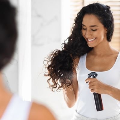 Smiling woman with curly hair, applying a protective hair formulation from a spray bottle to her ends.