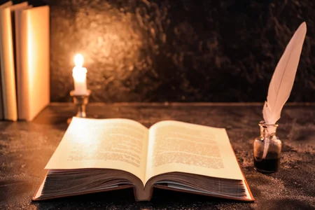Open old book with feather and burning candle on table